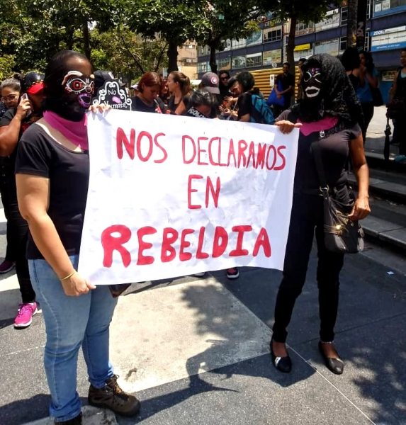 No meio da multidão, duas manifestantes encapuzadas seguram um cartaz. 