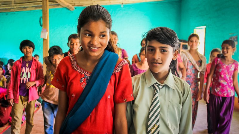 Deux jeunes enfants, une fille et un garçon, souriants et entourés par d'autres enfants, juste derrière eux. Ils se trouvent dans une salle de classe aux murs bleu turquoise.