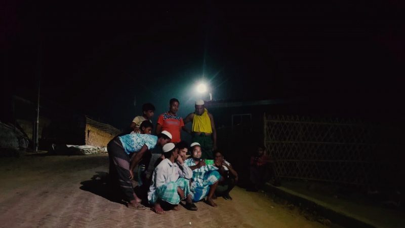 Photo de nuit, à la lumière d'un lampadaire. Un groupe d'hommes rohingya est regroupé autour de deux téléphones portables. Certains se penchent pour mieux voir.