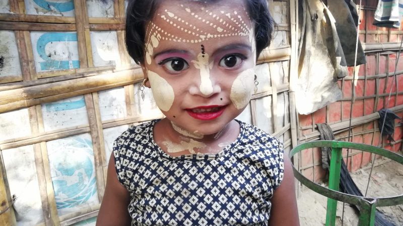 Une enfant maquillée et le visage orné d'un masque de chandan sourit à la caméra. Elle se tient devant un mur de bambou, colmaté avec une bâche de l'Unicef.