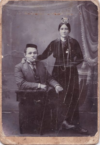 Une femme debout, portant un petit couvre-chef, tient par l'épaule un homme assis, vêtu d'un costume-cravate. La pose est solennelle.