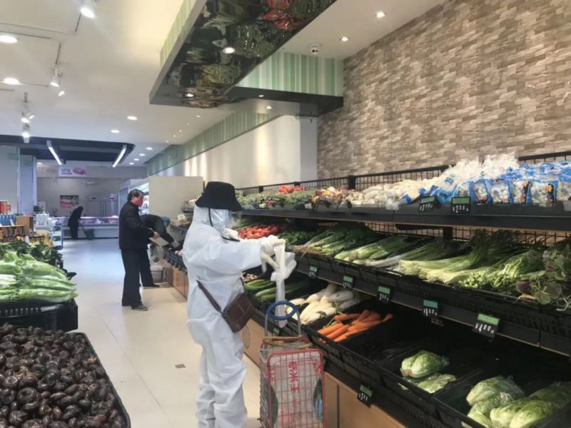 Quelques rares clients sont à l'intérieur d'un supermarché. Une personne, portant un équipement de protection complet avec un chapeau noir sur la tête, se trouve au rayon des fruits et légumes.