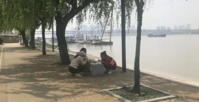 Trois femmes sont accroupies et piquent-niquent au bord d'une rivière, à l'ombre, sous les arbres. On distingue des bateaux, au loin.