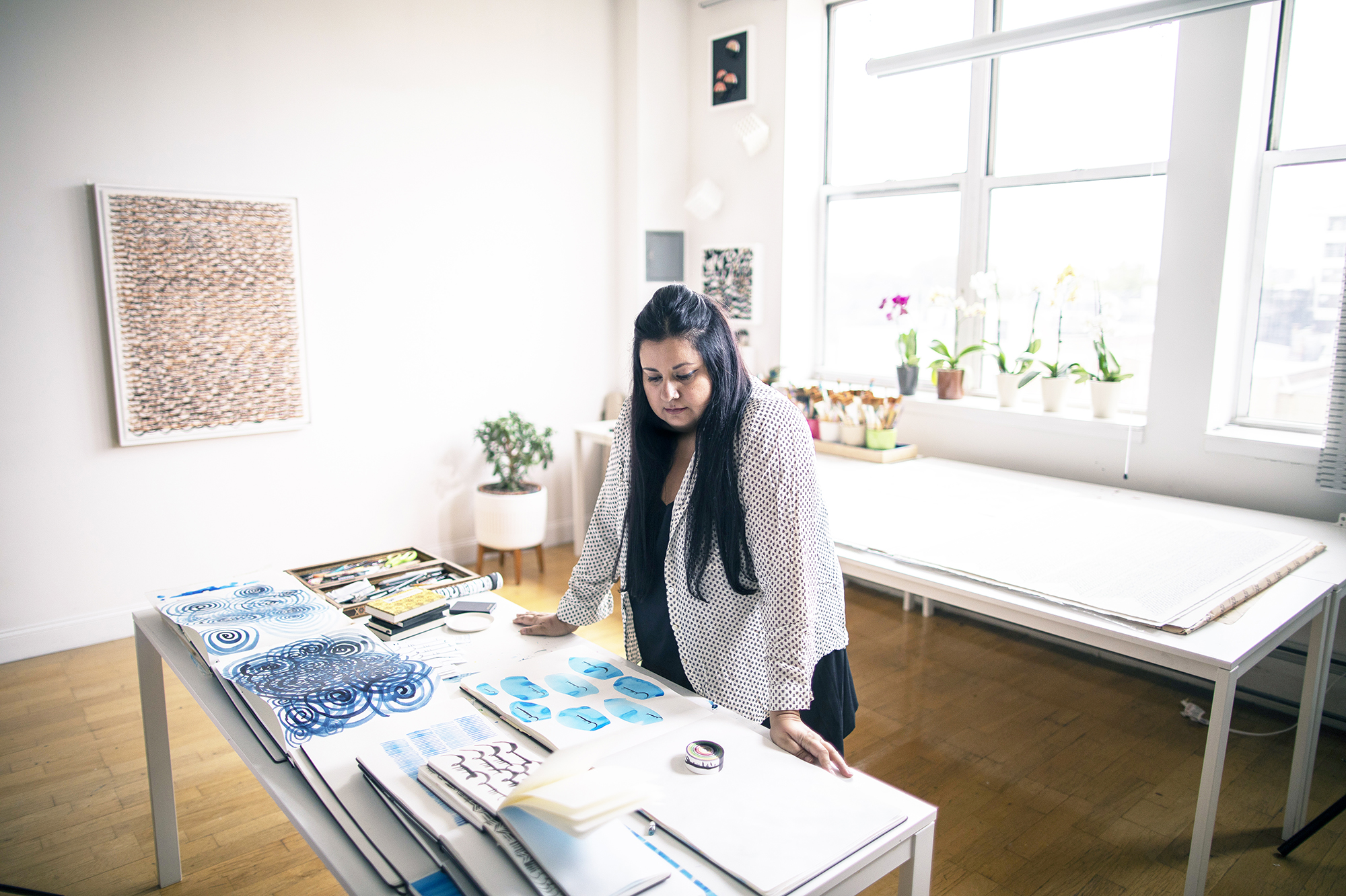 L'artiste dans son studio inondé de lumière. Elle a de longs cheveux bruns. Elle regarde quelques unes de ses créations dans des brochures, posées sur une table.