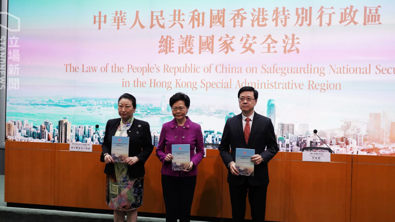 Deux femmes et un homme se tiennent debout, un document entre les mains, avec un air plutôt grave. En arrière plan, une grande affiche de Hong Kong. En gros caractères, en anglais et en chinois, on peut lire "La loi de la République populaire de Chine, sur la garantie du secret national, dans la région administrative spéciale de Hong Kong".