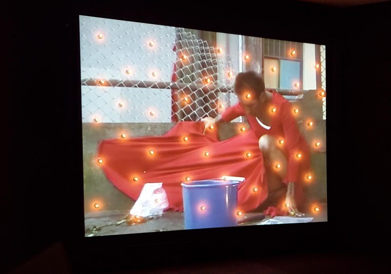 artiste Rebecca Belmore accroupie sur le sol, sa robe rouge clouée à un mur. Un seau est posé devant elle et des leds brillent sur toute l'image.