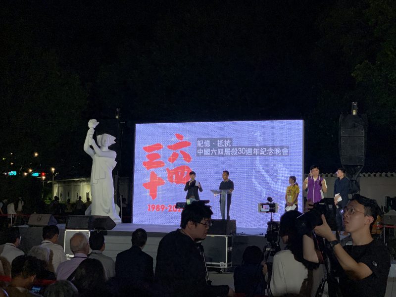 Sur une estrade se tient une statue blanche, portant un flambeau. Derrière, une grande affiche avec des inscriptions en chinois, et deux personnes avec un micro s'expriment devant un public.
