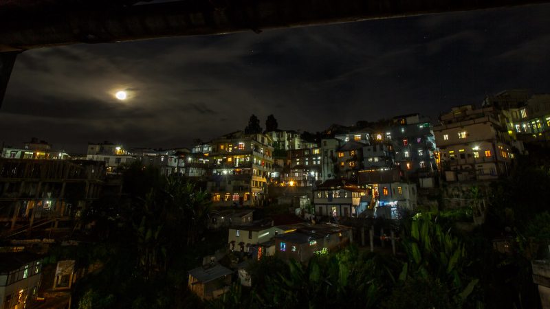 Image d'une ville en altitude, de nuit. On distingue de nombreuses maisons éclairées. Le ciel, occupant la moitié de l'image, est nuageux. La lune éclaire aussi la ville.