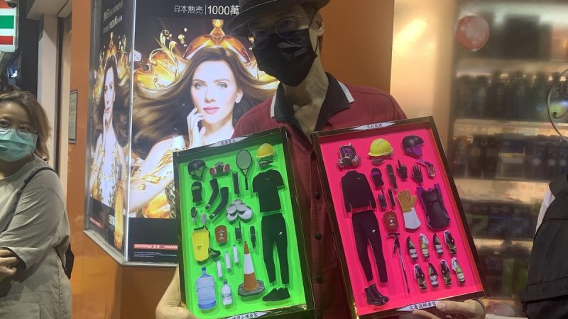 Un homme, chapoté, avec un masque de protection faciale, porte deux cadres où figurent tout l'équipement nécessaire pour manifester en toute sécurité (casque, masque à gaz...).