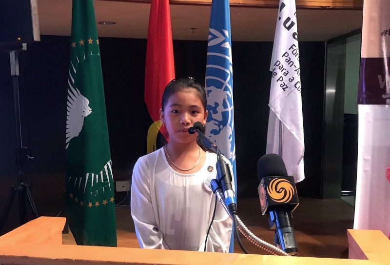 Licypriya se tient devant un micro, prête à prendre la parole. Derrière elle sont visibles les drapeaux de l'ONU et de l'Angola.