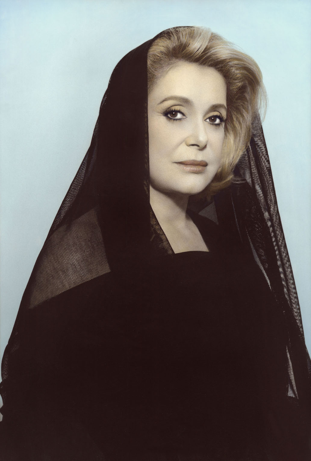 Image prise de l'actrice Catherine Deneuve, vêtue de noir et semi-voilée sur un arrière plan bleu ciel