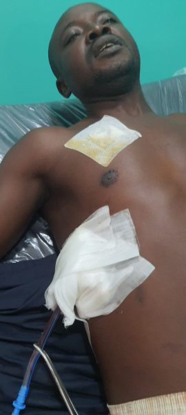 Nicholas Okpe est allongé, il semble souffrant. On voit un pansement sur sa poitrine et un autre sur son flanc droit.