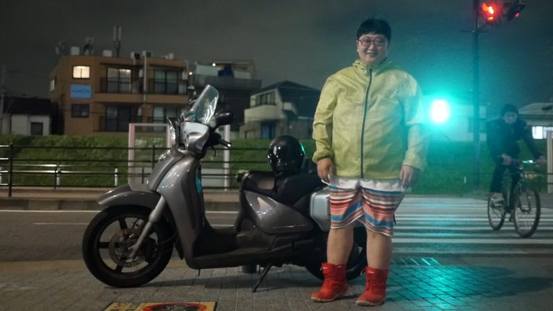 L'artiste Nogi Sumiko pris en photo de nuit devant un scooter. Il porte un imperméable, un short rayé et des bottines rouges.