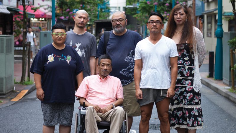 Six personnes figurant dans le documentaire Queer Japan. Cinq d'entre eux sont debout, affichant une expression neutre et vêtus d'habits légers. Le sixième est un vieil homme assis devant eux dans son fauteuil roulant.