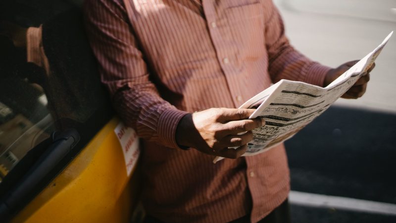 L'image montre un homme, sans montrer sa tête, tenir un journal entre ses mains et le lire. Il est adossé à une voiture jaune, vraisemblablement un taxi. Il porte une chemise rayée de couleur vieux rose. On aperçoit la lumière des rayons du soleil sur l'une de ses mains.