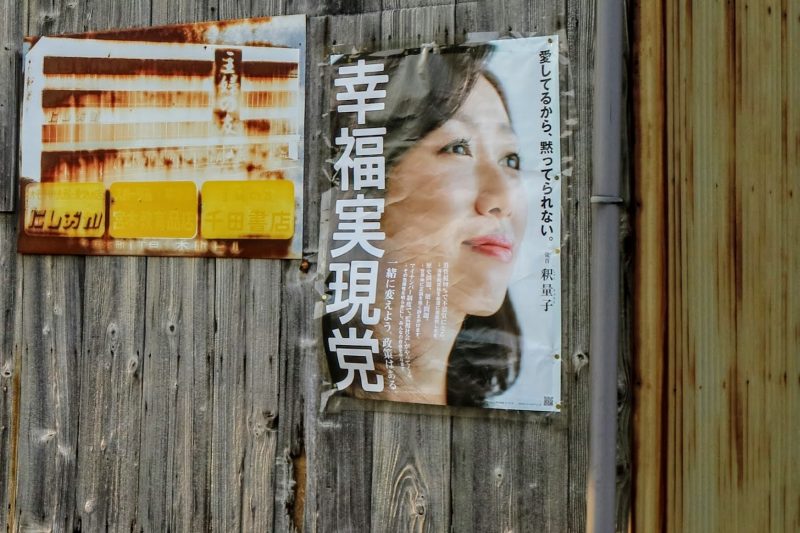 Il s'agit d'une affiche, représentant le « Parti de la réalisation du bonheur » (Happiness Realization Party). On y voit le profil d'une femme avec un visage plutôt souriant, collée sur un mur de bois. On lit des caractères en japonais.