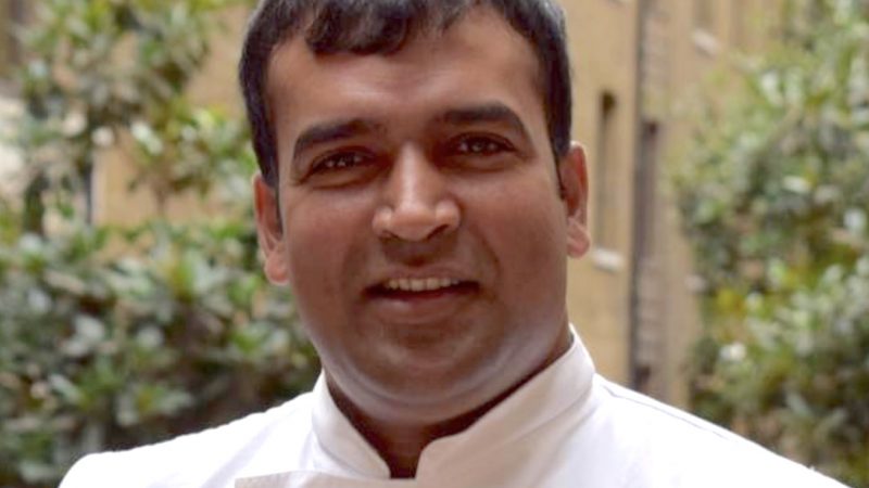 Le chef cuisinier Santosh Shah arbore un demi-sourire. Il a des joues rebondies et porte une blouse blanche.