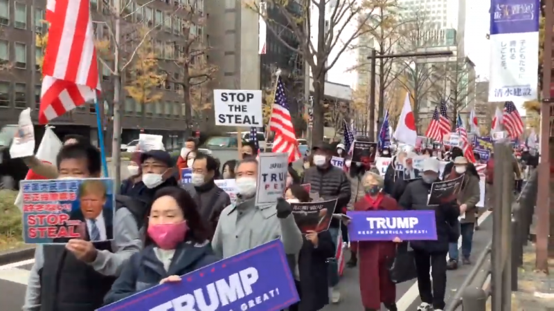 Manifestations de nombreuses personnes, portant un masque de protection faciale, au Japon. Elles marchent avec des drapeaux américains et japonais. Il y a aussi différentes banderoles en faveur du président américain sortant, Donald Trump : « stop the steal » (Stoppez l'escroquerie), en japonais et en anglais, « keep America great ! » (Maintenons la grandeur de l'Amérique). Le défilé se tient dans une rue encadrée de hauts buildings, en journée.