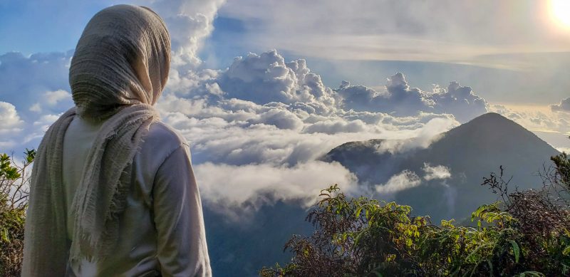Жінка зі спини із хусткою на голові, яка дивиться на хмарний пейзаж.