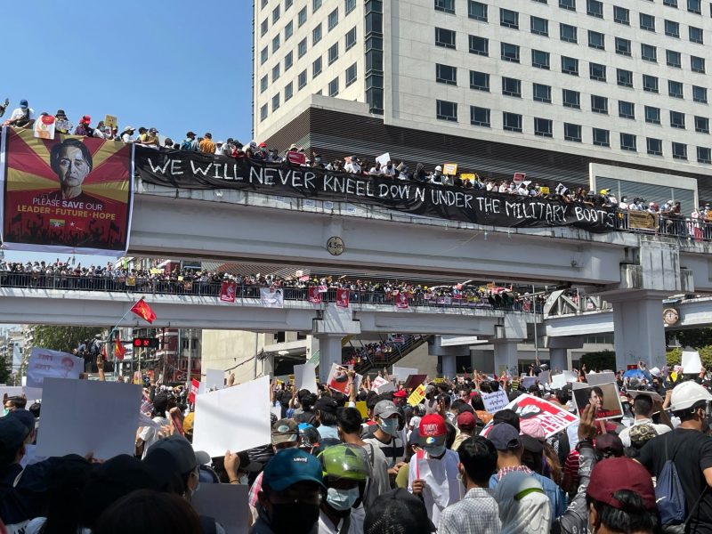 L'image montre une foule compacte de manifestants dans la rue et sur des passerelles. Nombreux sont ceux qui brandissent des pancartes avec des revendications. Sur la 1ère passerelle, une banderole noire est tendue avec l'inscription en anglais avec une couleur blanche : "We will never kneel down under the military boots" (Nous ne nous agenouillerons jamais sous les bottes des militaires). A gauche de cette banderole, on voit une affiche avec le visage de Aung San Suu Kyi. La scène se passe en journée, sous le soleil. En arrière plan, on distingue un building et un ciel bleu.
