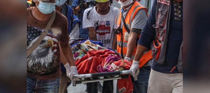 L'image montre plusieurs secouristes transporter un civil blessé par les tirs des forces de sécurité. Le blessé est sur une civière, enveloppé dans des couvertures. Le logo du site « The Irrawaddy » est visible sur le haut de l'image à gauche.