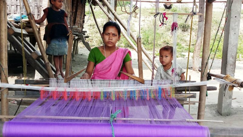 Priyanka Pegu from Bholukaguri village in Assam’s Golaghat district weaves a Mekhla chador in the backyard of her home, while her son looks on. Image Credit: NEADS. Used with permission.