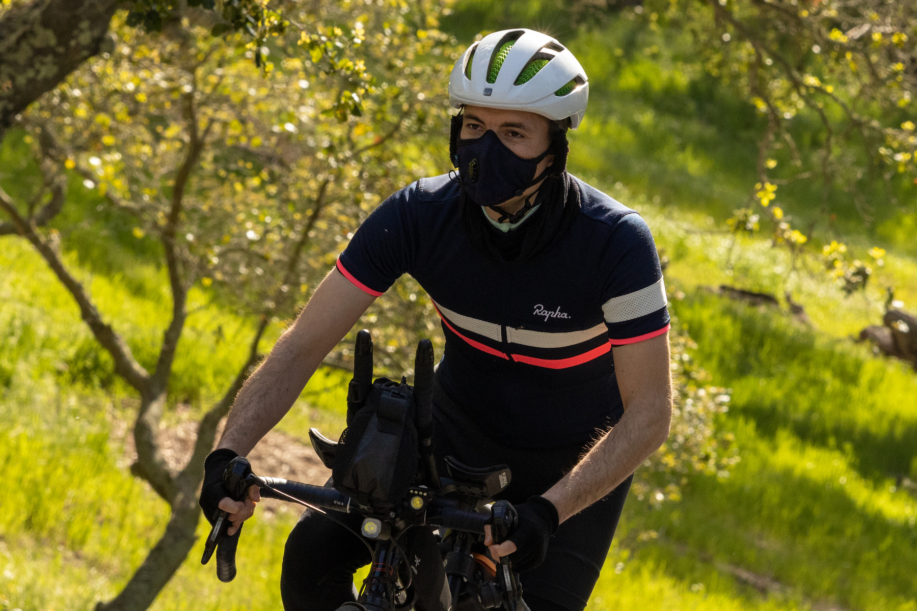 A cyclist wearing a helmet and mask steps out of the seat to climb a hill, with green trees in the background.